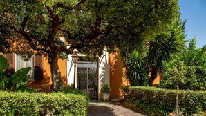 Un chemin menant à un bâtiment doté d’une grille en métal, entouré d’une végétation luxuriante et de plantes en pot.
