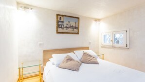 Ein Schlafzimmer mit einem hölzernen Kopfende, einem Bett mit weißen Leinen und zwei beigen Kissen, einem Nachttisch aus Glas, einem gerahmten Gemälde an der Wand und einem Fenster mit Blick auf ein Gebäude.