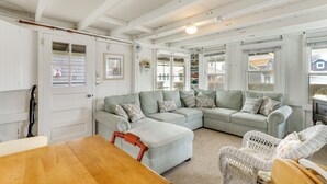 A cozy living room with a light blue sectional sofa, a wooden dining table, wicker chairs, and large windows offering a view of the outside.