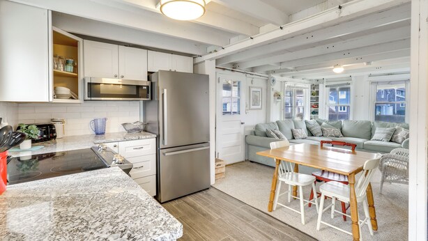 A modern kitchen with white cabinets, a stainless steel refrigerator, and a microwave. There is a dining area with a wooden table and white chairs.
