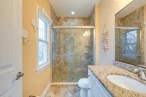 Un baño con ducha de vidrio, inodoro blanco, lavamanos con encimera de granito y un espejo grande.
