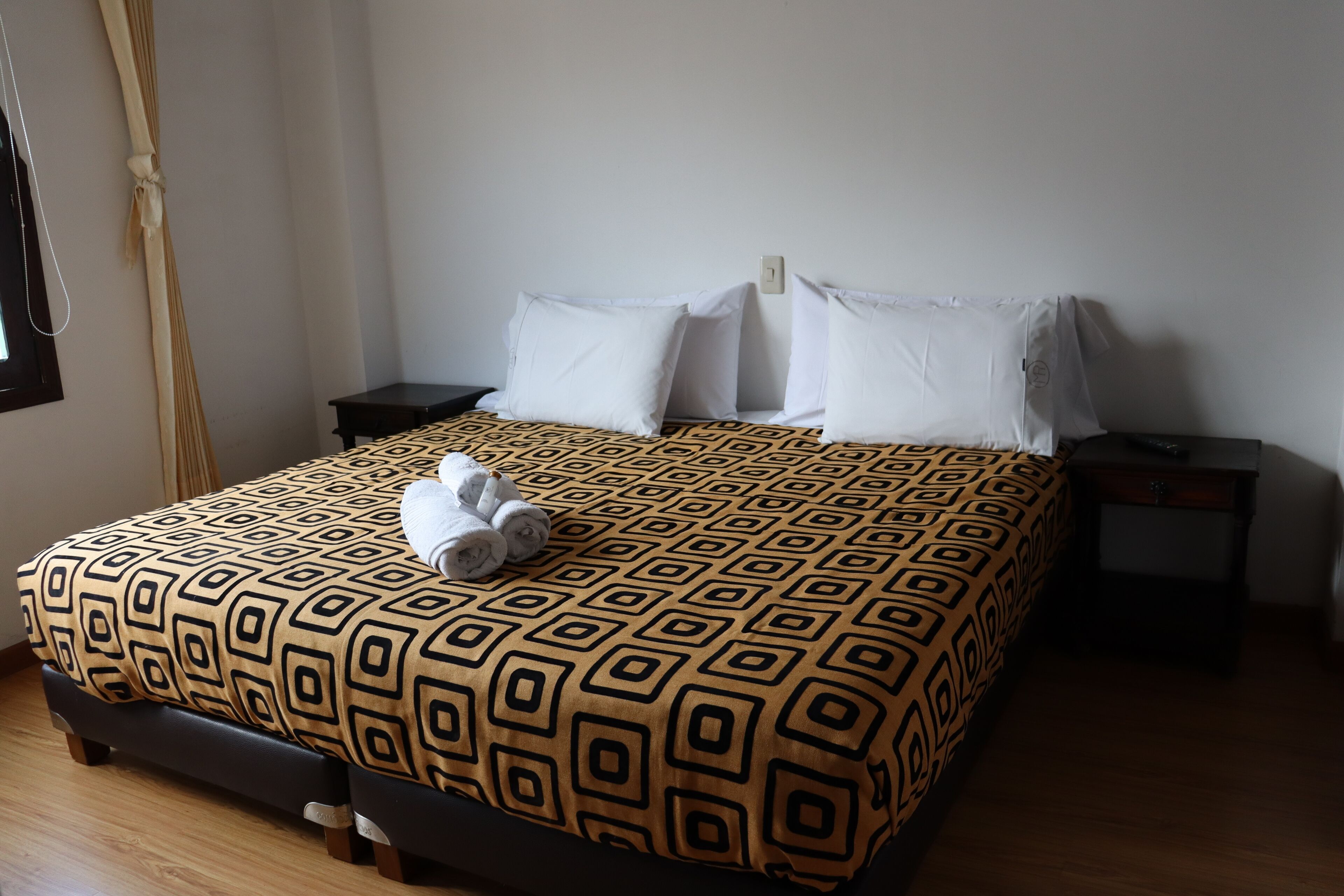 A neatly made bed with a geometric patterned bedspread, two bedside tables, and a window with curtains.