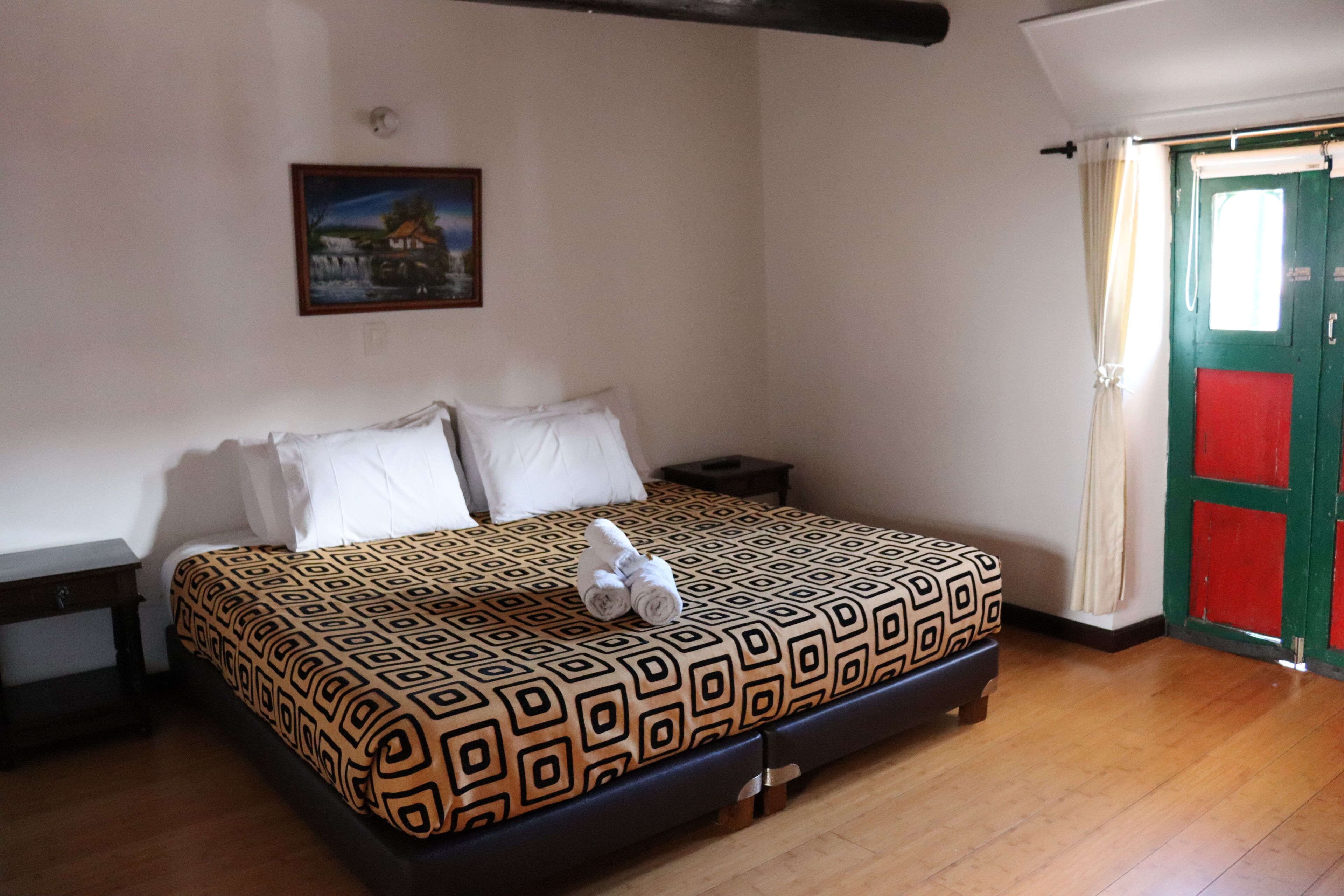 Un dormitorio con una colcha estampada, una mesita de noche, una pequeña mesa, un cuadro en la pared y una puerta con un panel rojo.