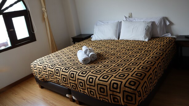 Een net opgemaakt bed met een bedsprei met dessin, een paar opgerolde handdoeken en een raam met gordijnen.