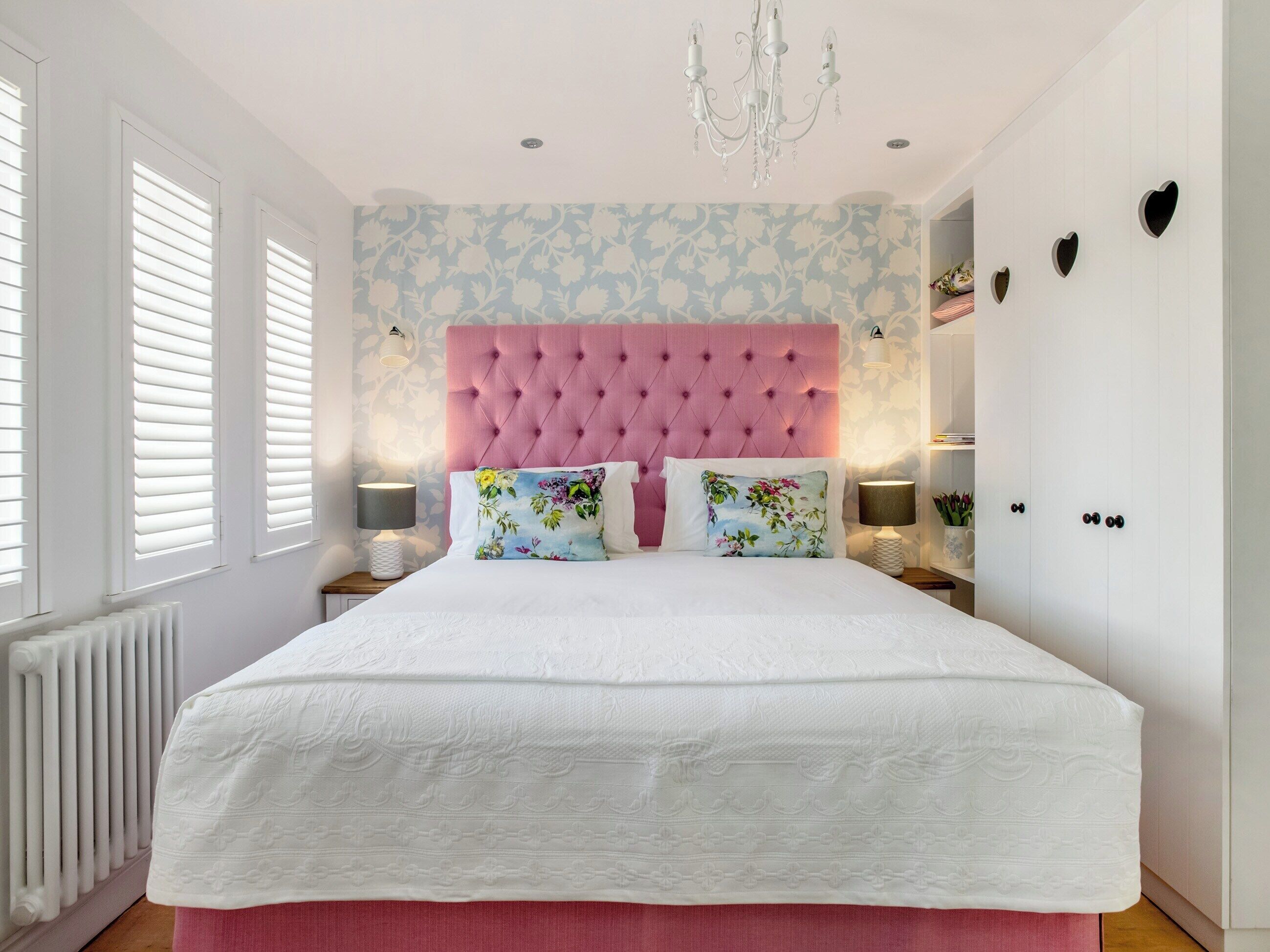 A bedroom with a pink tufted headboard, floral patterned wallpaper, a white bedspread, and two bedside lamps.