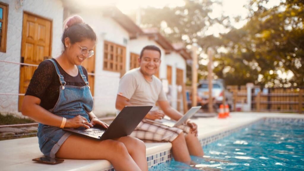 Two people sitting by a poolside, each using a laptop.