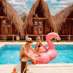 En swimmingpool med en rosa uppblåsbar flamingofigur som flyter i vattnet, omgiven av byggnader med halmtak.