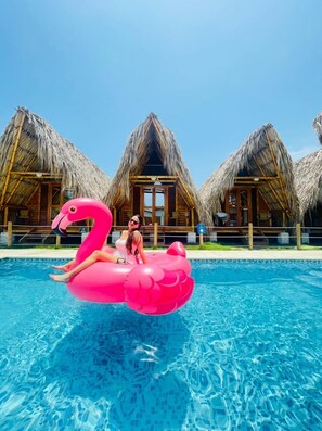 A swimming pool with a person on a pink flamingo float, surrounded by thatched-roof huts. - Palomino Sunrise (Dibulla)