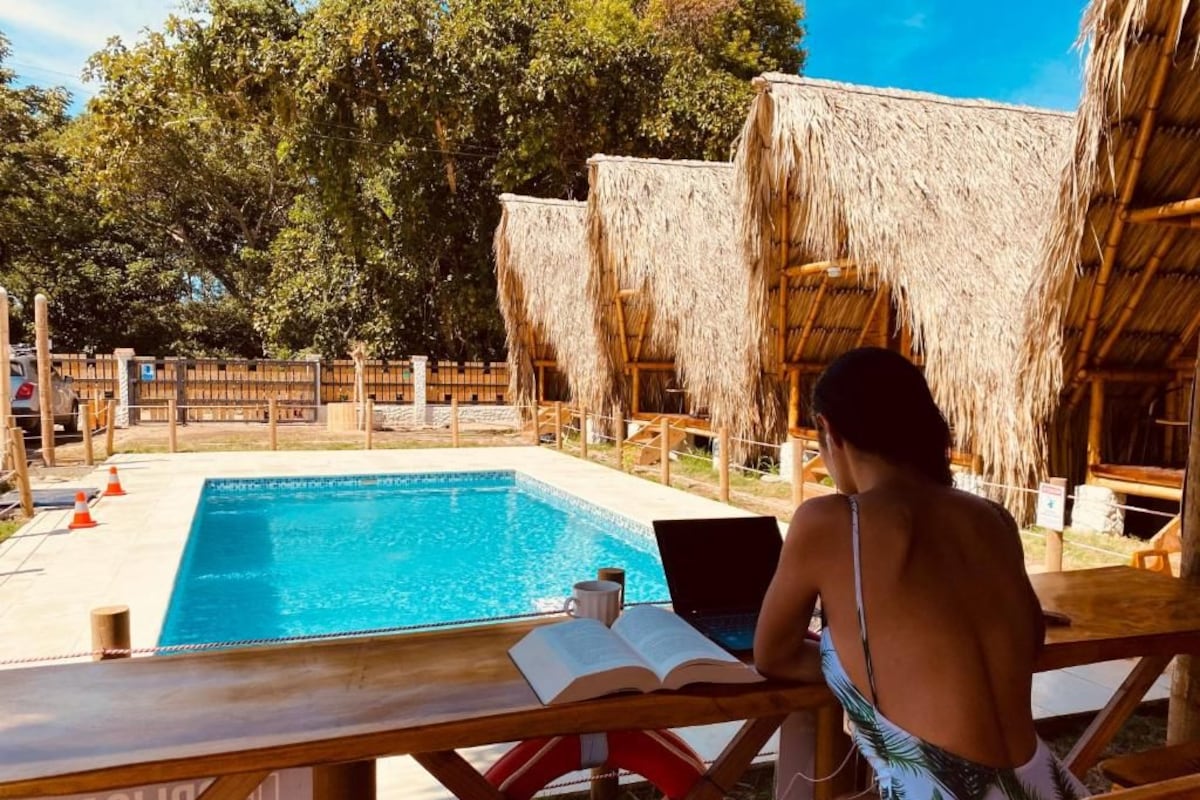 A person reading a book by a pool with thatched-roof structures in the background.