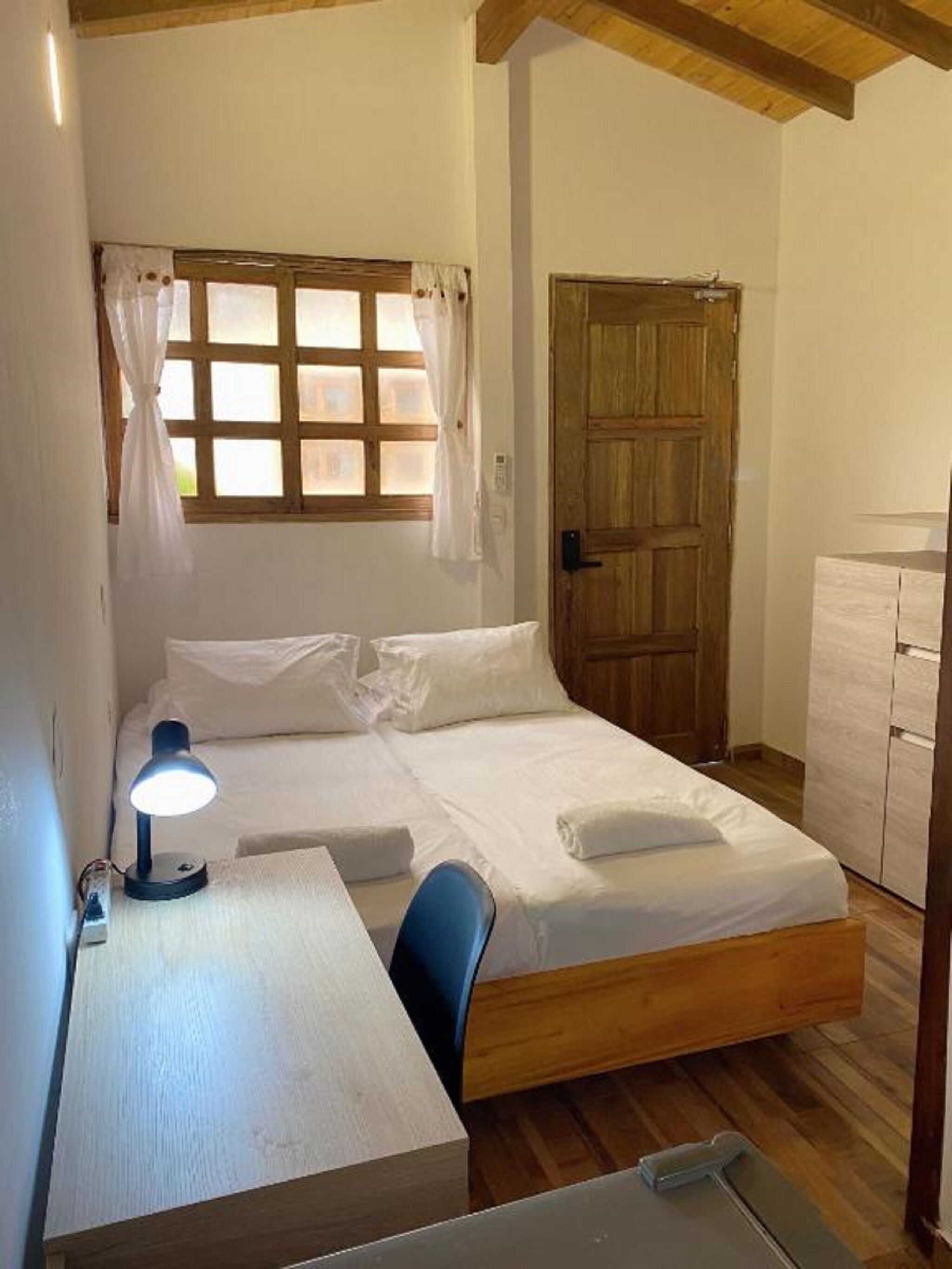 Un letto singolo con testiera in legno, un comodino con una lampada, una finestra con tende e una porta in legno.