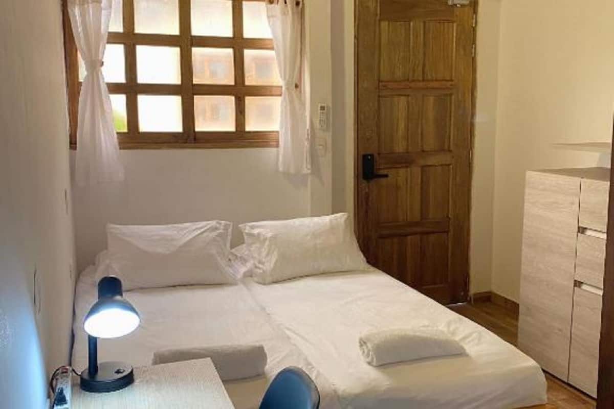 Ein Einzelbett mit Holz Kopfteil, ein Nachttisch mit Lampe, ein Fenster mit Vorhängen und eine Holztür.