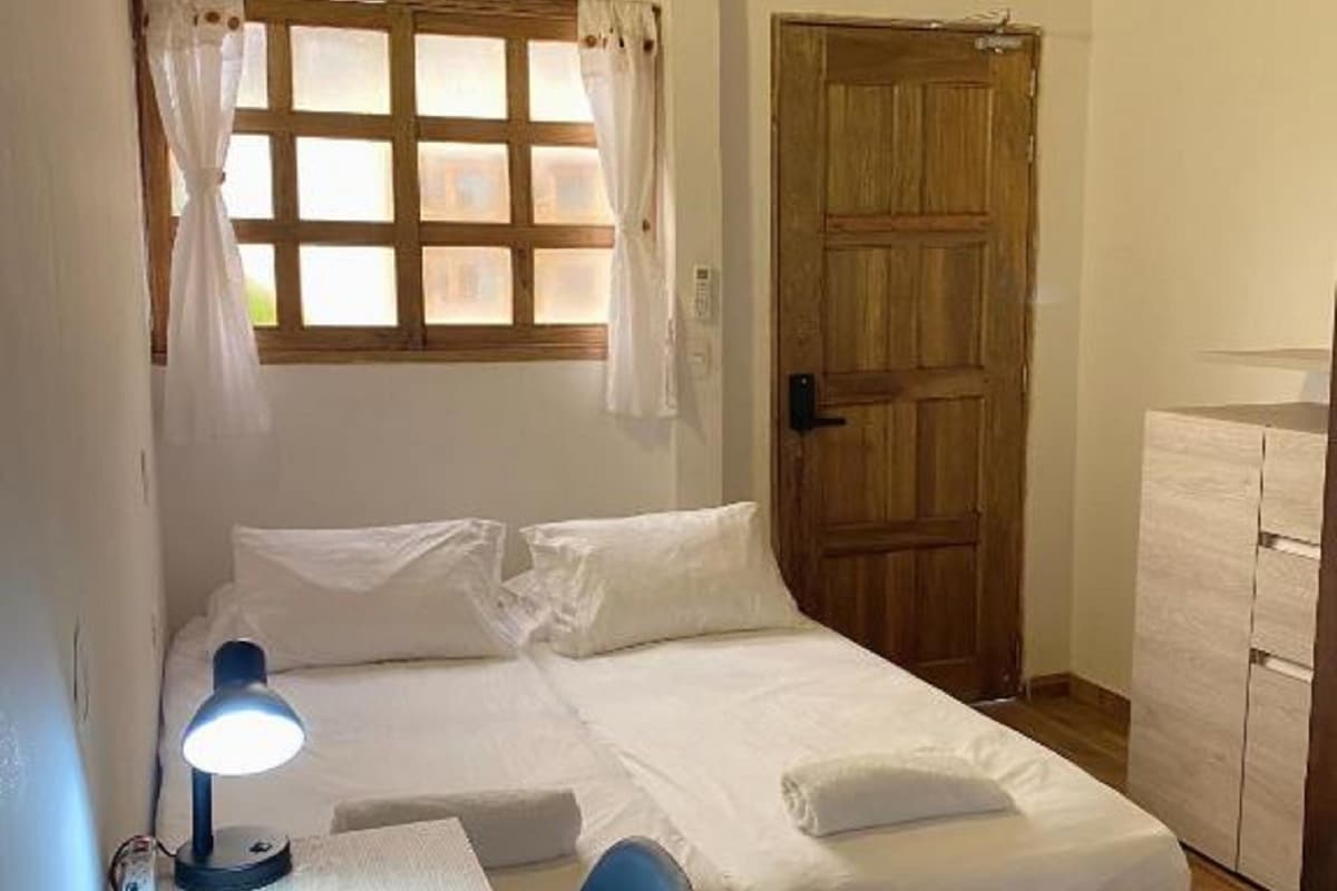 Una cama individual con cabecera de madera, una mesita de noche con lámpara, una ventana con cortinas y una puerta de madera.