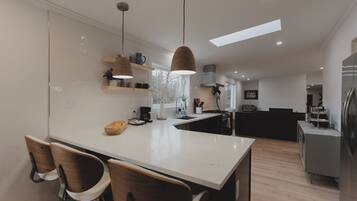 Une cuisine moderne avec des plans de travail blancs, des chaises en bois, des luminaires suspendus et un four encastré.