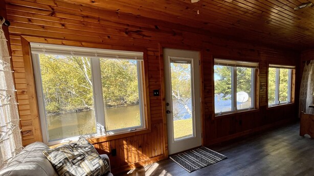 A cabin interior with a wooden floor, large windows, a couch, and a door leading outside. - Riverfront Retreat | Fire, Sunroom, Kayaks, Tubes (Sears)