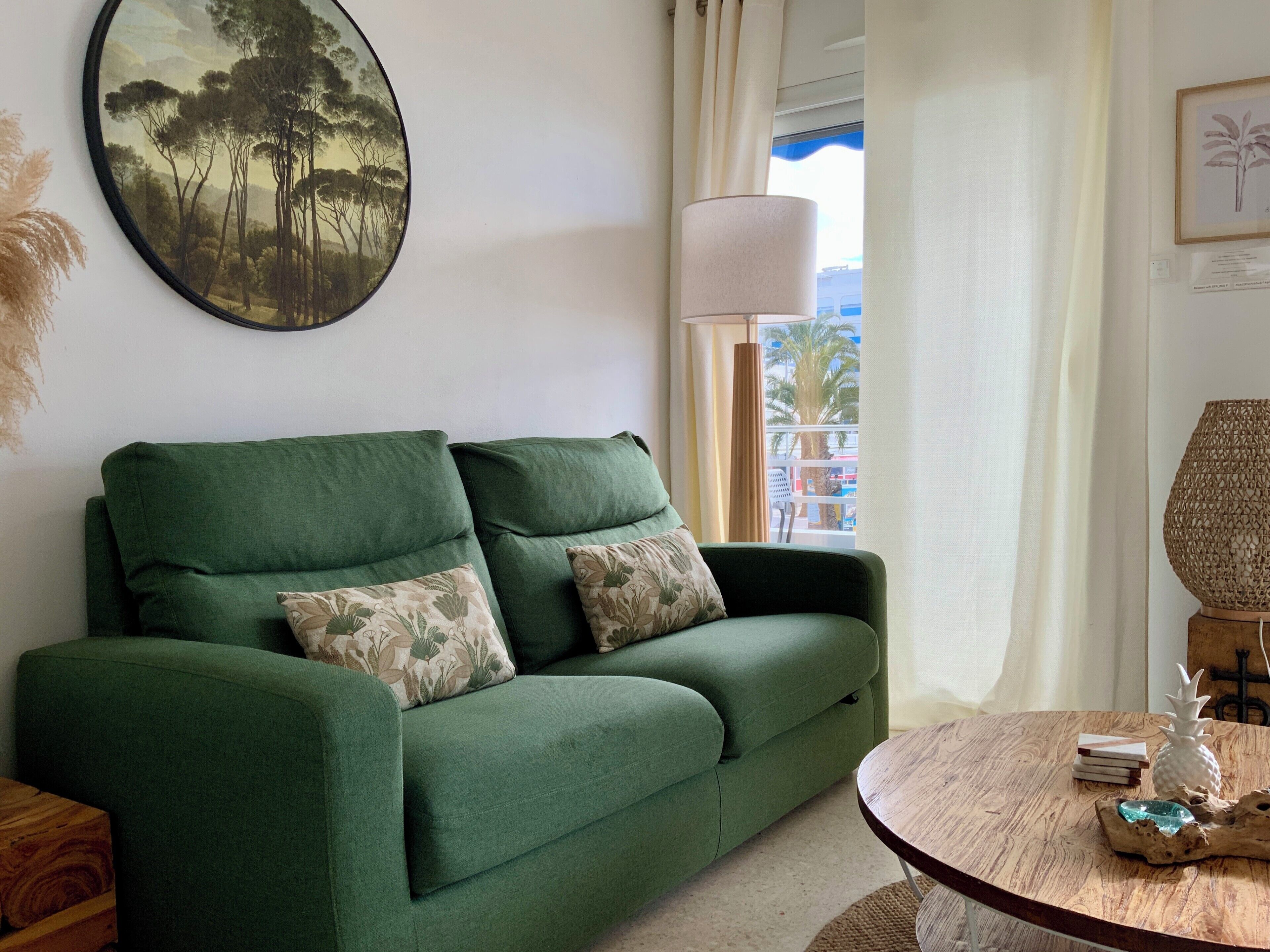 Un sofá verde con cojines estampados, una mesa redonda de madera con objetos decorativos, un cuadro grande enmarcado y una ventana con vista a palmeras.