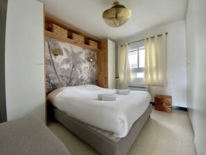 A neatly made bed with white linens and two towels, a wooden headboard with woven baskets, a ceiling light fixture, and a window with curtains. - Impérial 1 – Appartement vue mer face à la plage Rive Gauche du Grau-du-Roi (Le Grau-du-Roi)