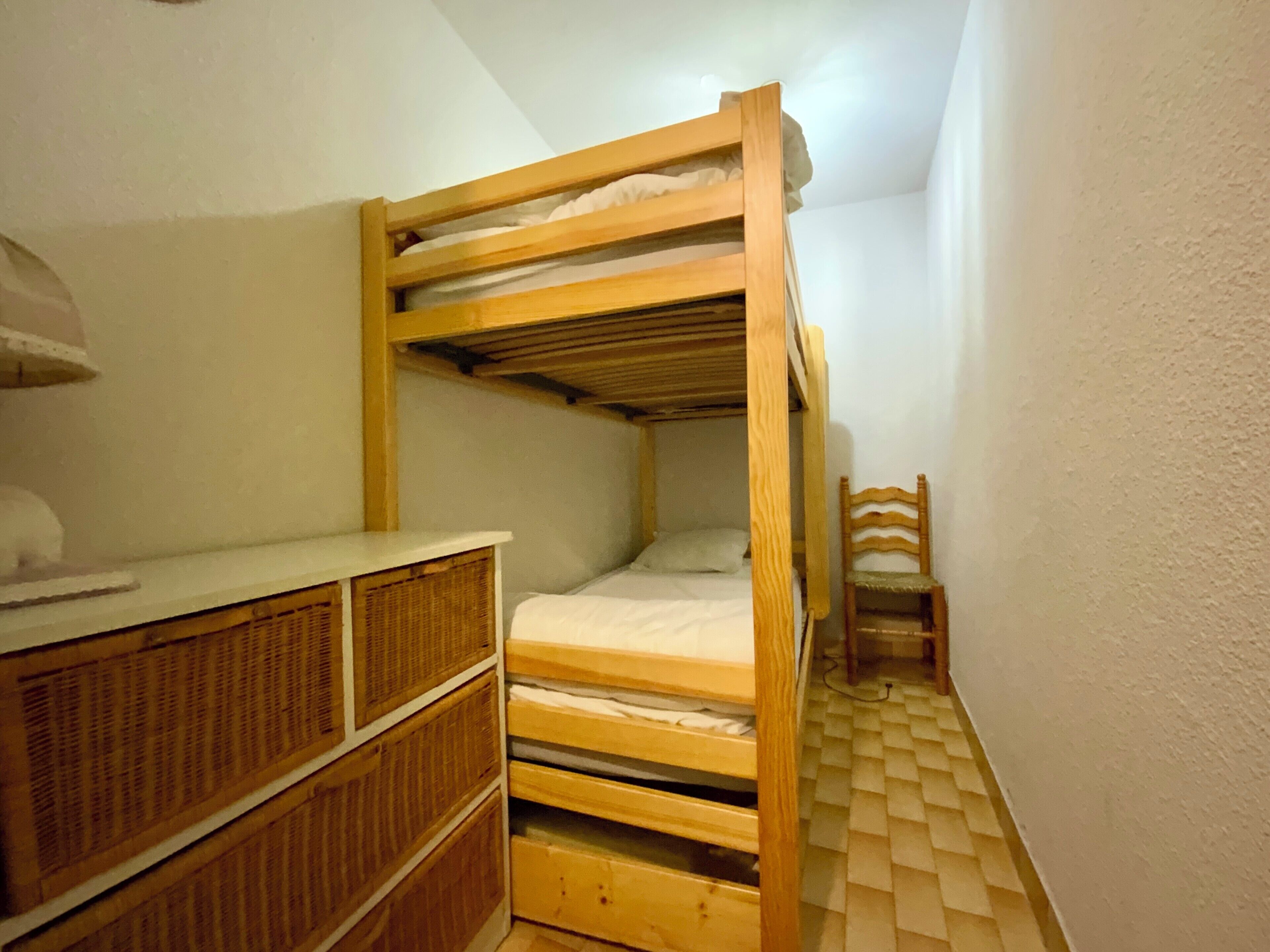 A small, narrow room with a bunk bed, a wooden dresser, and a chair.