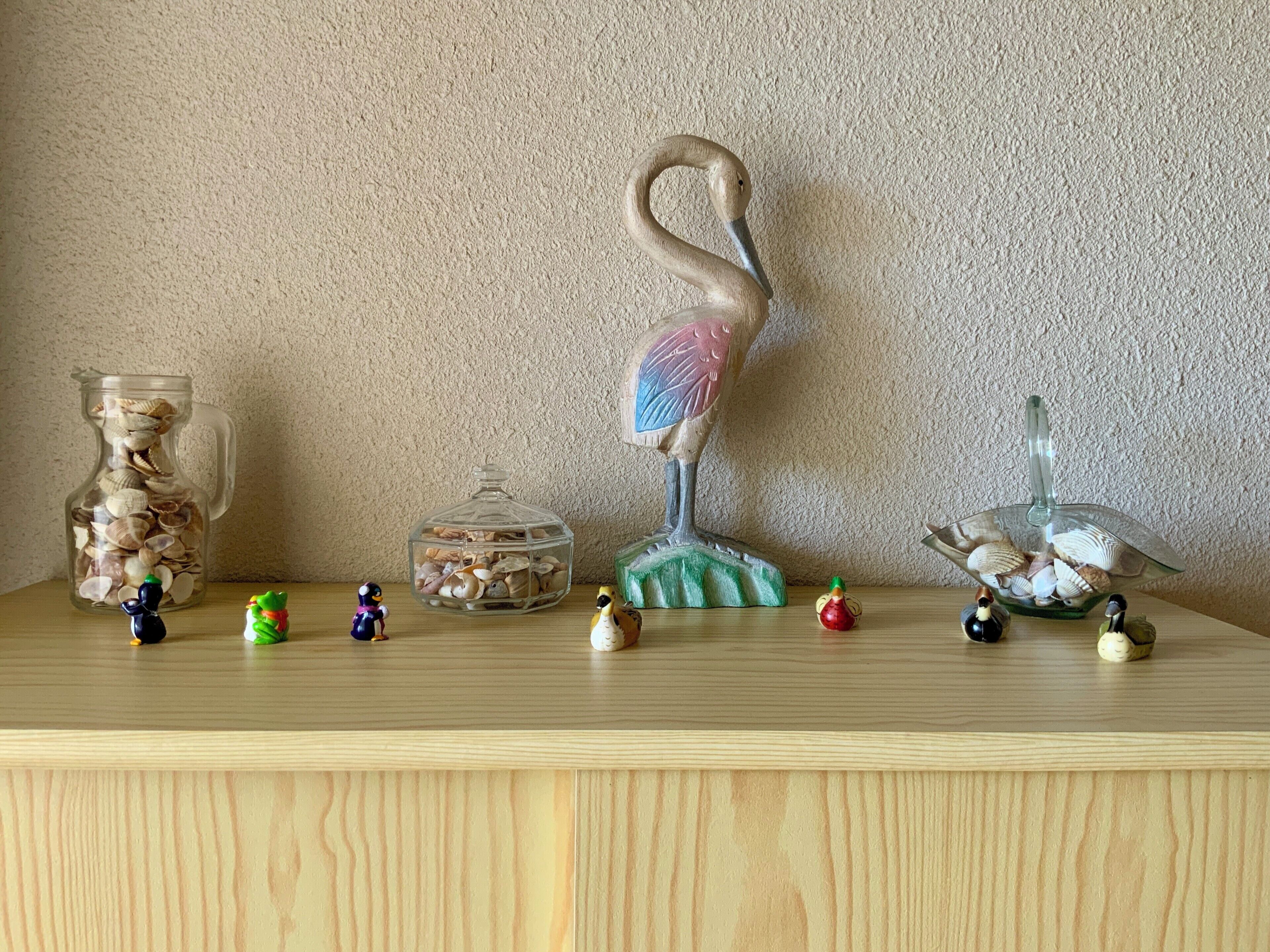 A wooden shelf with various decorative items including a glass pitcher filled with seashells, a glass birdhouse, and several small figurines.