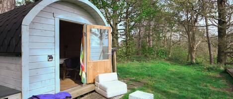 A small, single-room cabin with a door open, a white bench outside, and a picnic table with a basket.