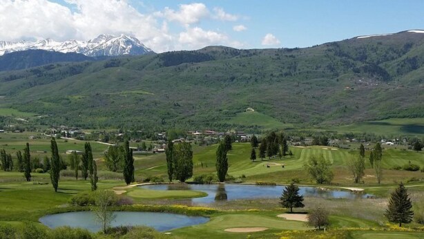 Ein Golfplatz mit Teichen und Bäumen, umgeben von Bergen und einer kleinen Stadt.