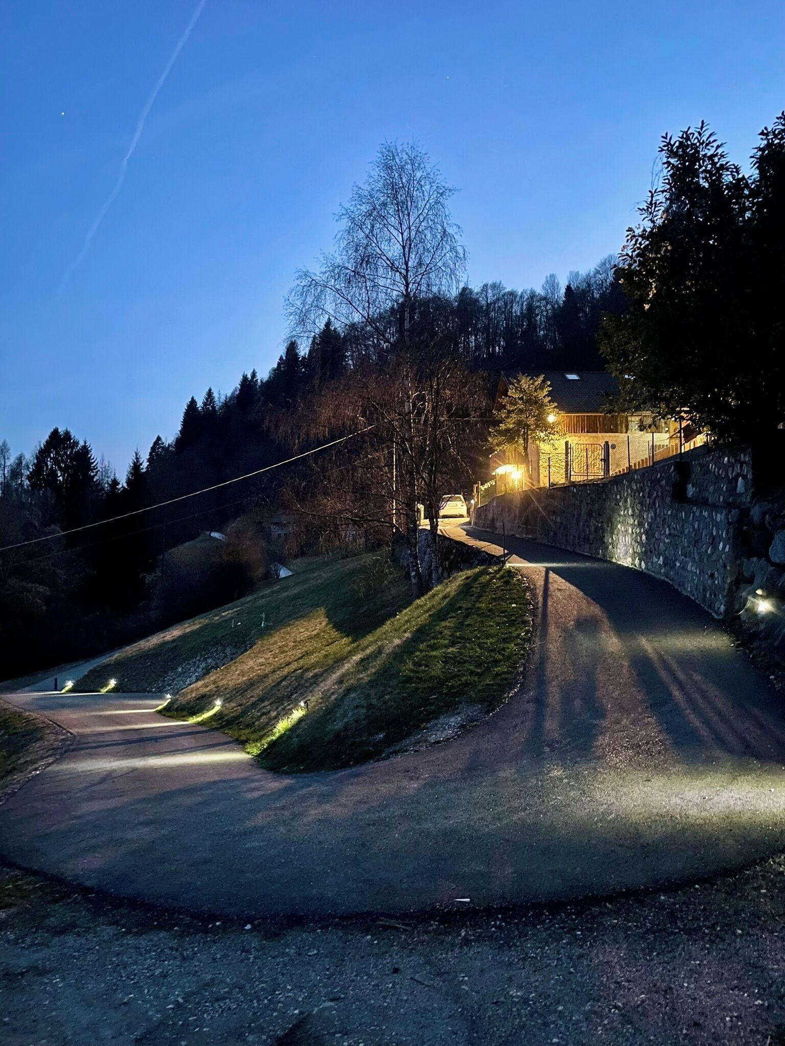 Un camino rural que lleva a un edificio de paredes de piedra con una entrada iluminada, rodeado de árboles y colinas.