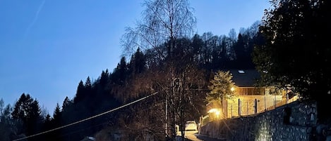 Un camino rural que lleva a un edificio de paredes de piedra con una entrada iluminada, rodeado de árboles y colinas.