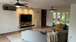 A modern living room with a curved sofa, a flat-screen TV, ceiling fans, and a large window with a view of greenery. - Hidden Paradise w/ Pool (Guaynabo)