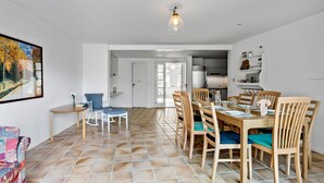 Uma sala de jantar com mesa e cadeiras de madeira, um quadro na parede e uma cozinha ao fundo.