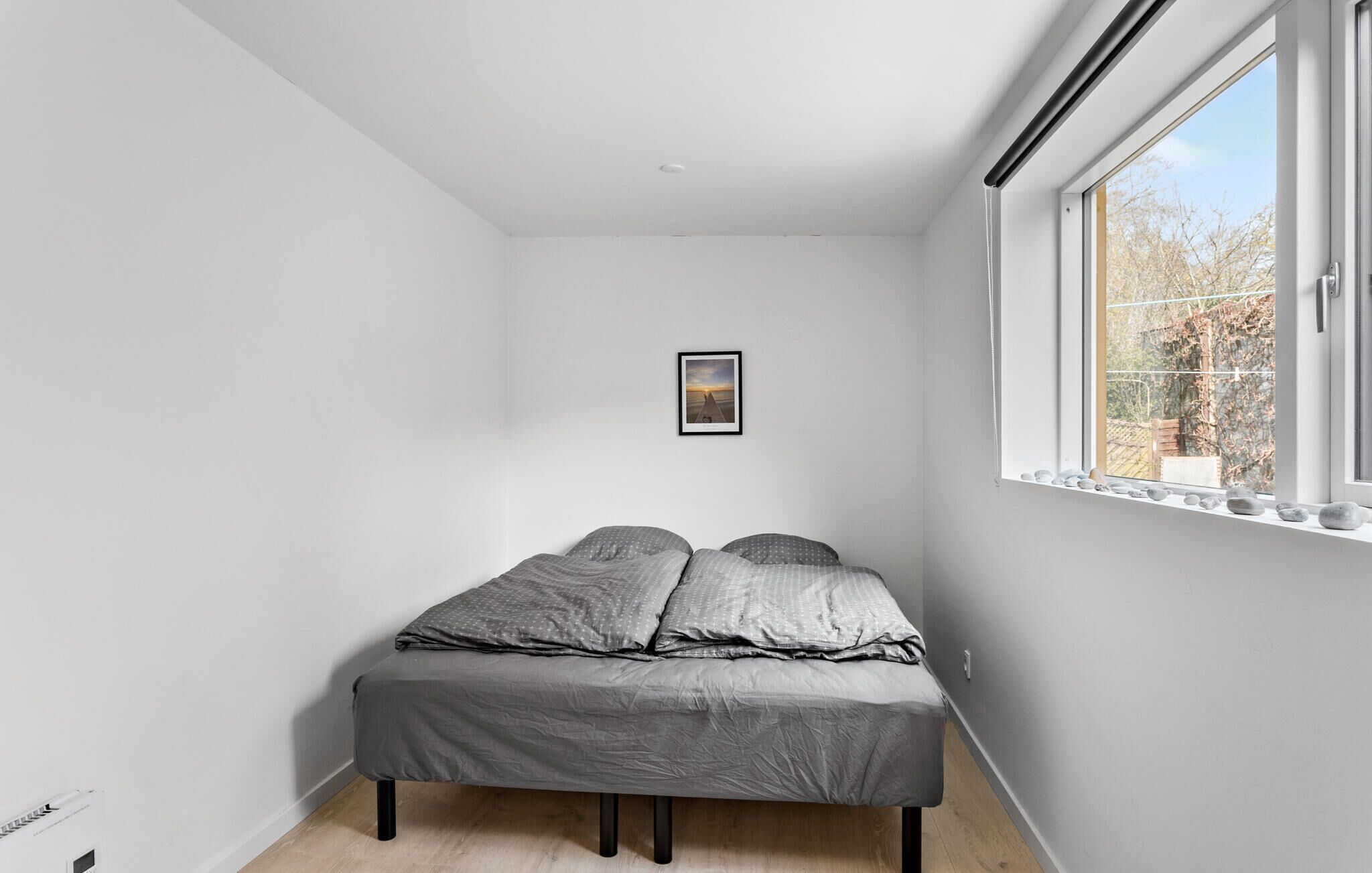 A small, bright room with a bed, a framed picture on the wall, and a window with a view of trees.
