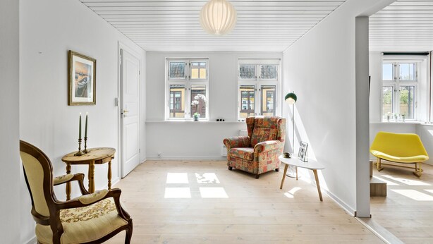 Quarto espaçoso com piso de madeira, poltrona estampada, cadeira amarela e uma mesa com luminária.