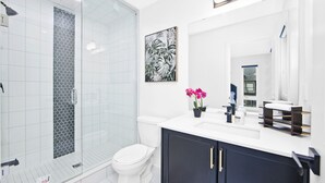 Une salle de bain moderne avec une douche à l’italienne en verre, des toilettes blanches, un meuble-lavabo foncé avec un miroir et une orchidée en pot.