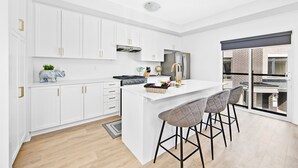 Une cuisine moderne avec des meubles blancs, un îlot central doté de tabourets, un réfrigérateur en acier inoxydable et une grande fenêtre offrant une vue sur la ville.