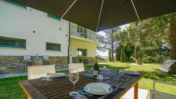 An outdoor dining area with a wooden table set for two, a bottle of wine, and a glass. The setting includes a stone wall, a balcony, and a view of a lush garden. - Cozy home in Marsaglia with kitchen (Marsaglia)