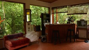 Una habitación acogedora con un sofá rojo, una zona de bar de madera y amplios ventanales con vistas a exuberante vegetación.