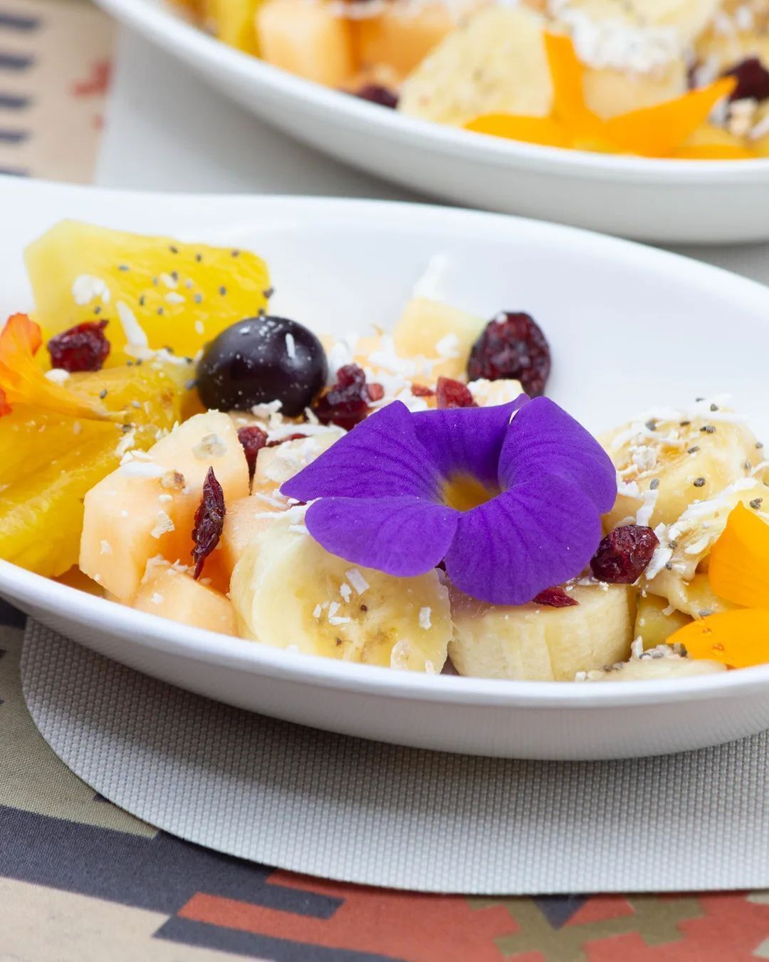 Une assiette blanche contenant une salade de fruits, notamment des bananes, des oranges et une fleur violette en guise de décoration.