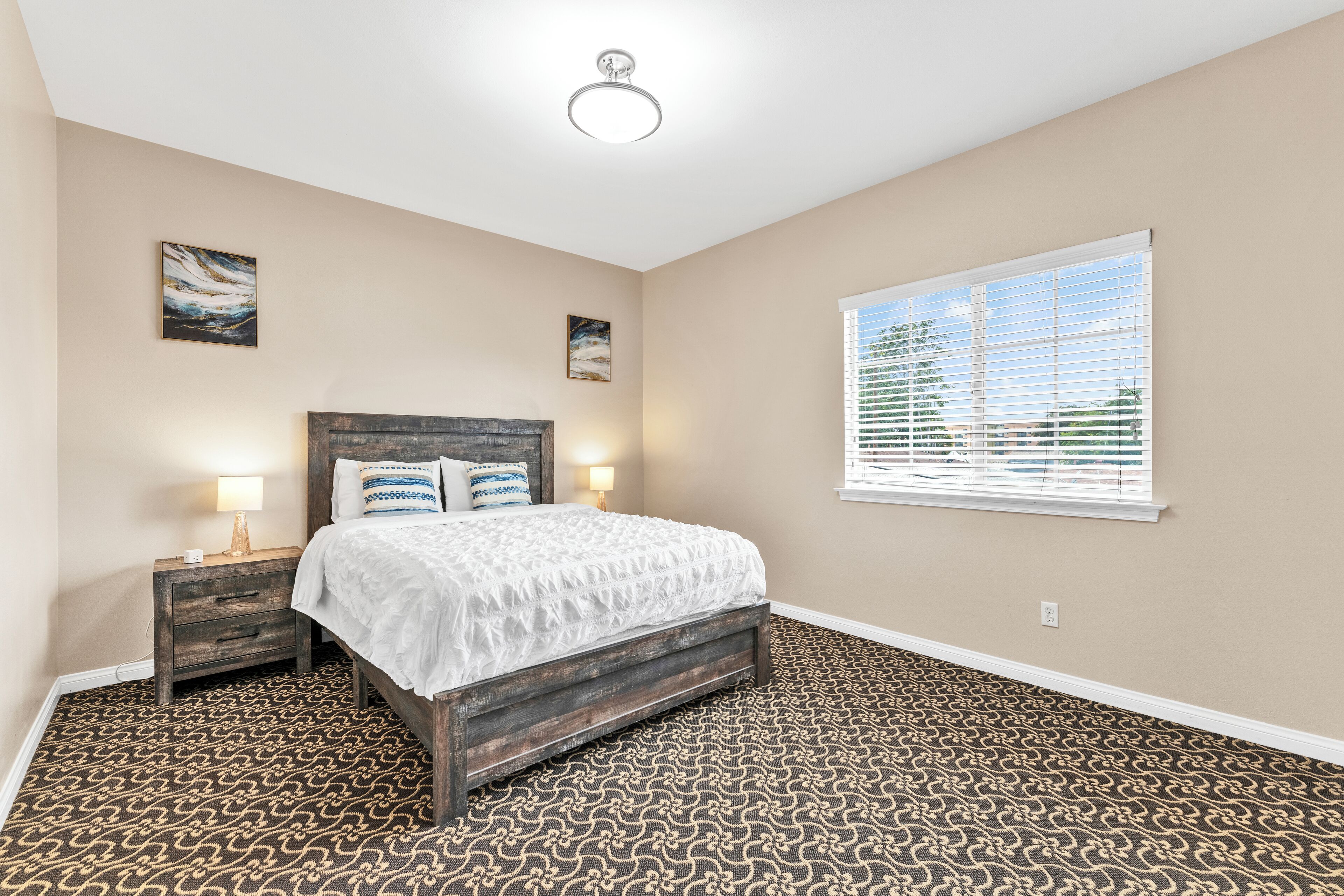 A bedroom with a patterned carpet, a wooden bed frame, two bedside tables with lamps, and a window with blinds.
