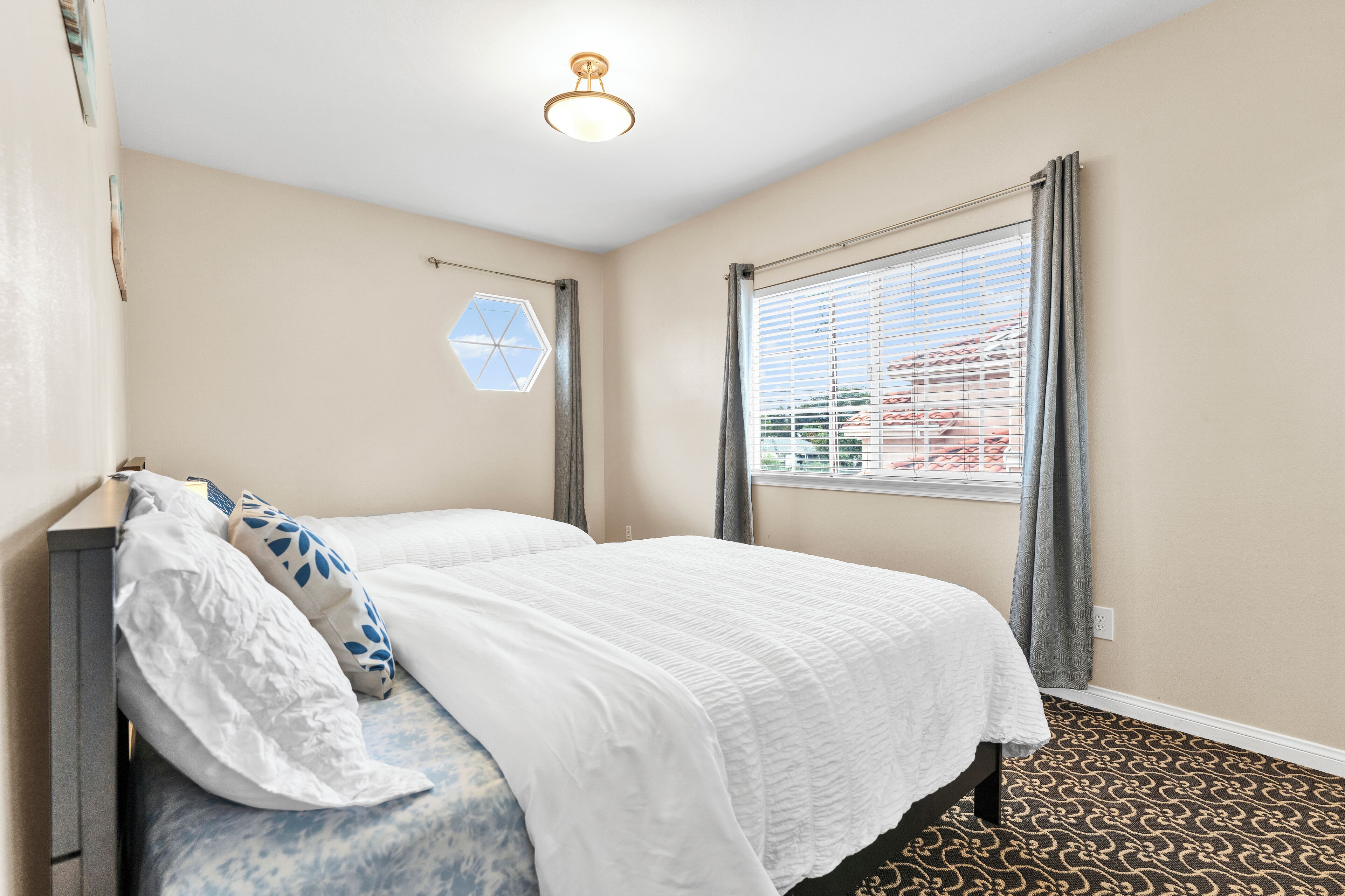 A neatly made bed with white bedding and a patterned headboard, a window with blinds, and a hexagonal light fixture on the wall.