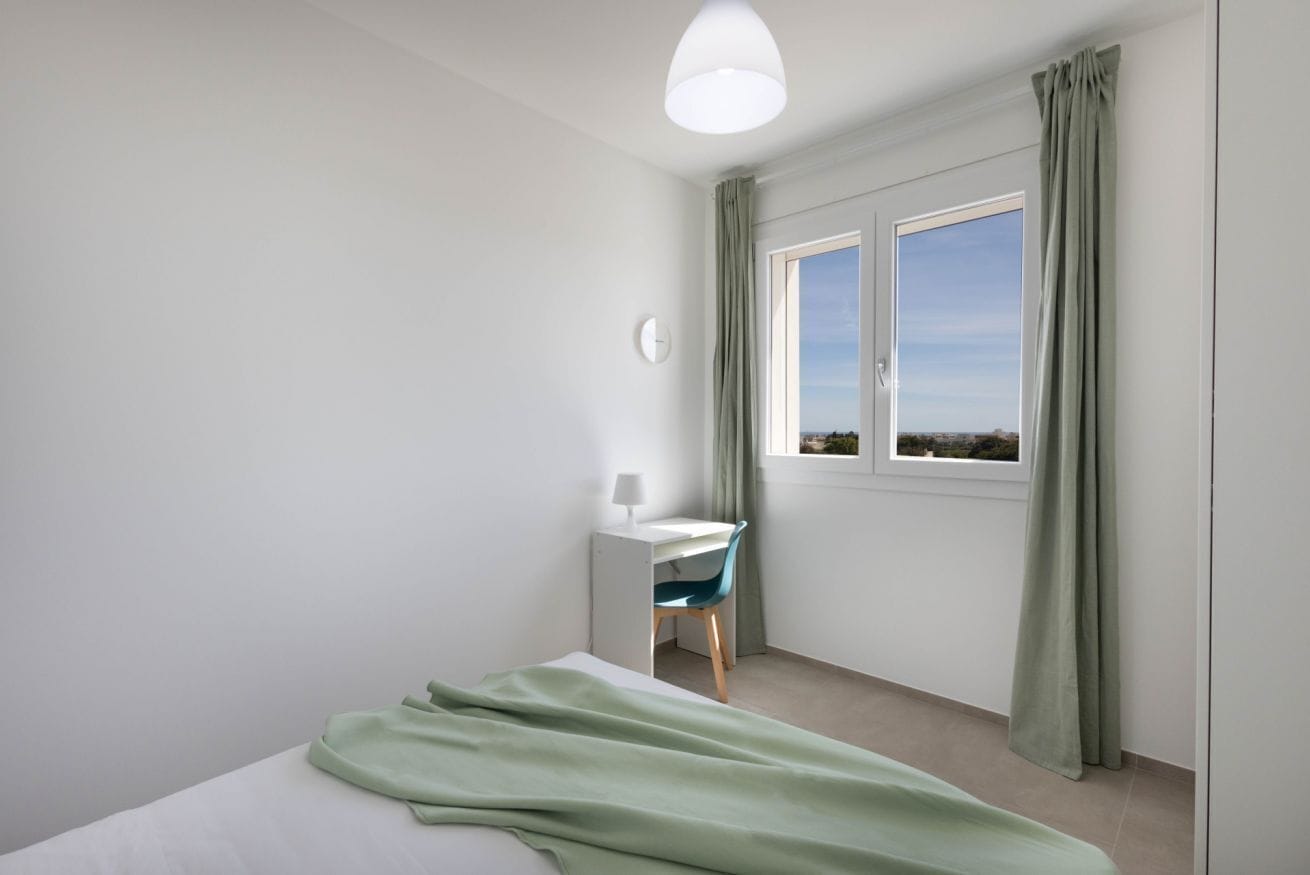 Une chambre à coucher avec un lit, un bureau, une chaise, une fenêtre avec des rideaux et une horloge au mur.