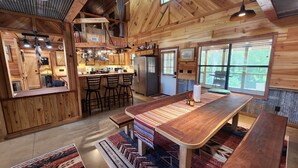 L’intérieur d’un chalet rustique comprend une table à manger en bois, des tabourets de bar, un coin cuisine et une grande fenêtre offrant une vue sur l’extérieur.