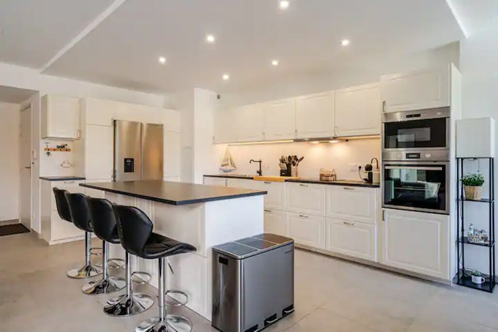 A modern kitchen with white cabinets, a central island with bar stools, stainless steel appliances, and recessed ceiling lighting.