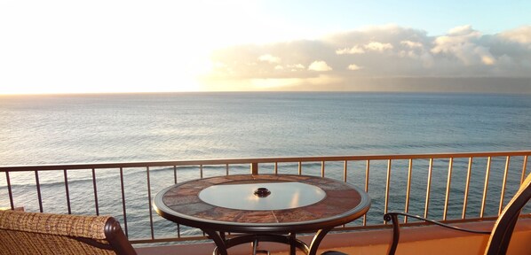 Ein Balkon mit rundem Tisch und einem Stuhl, Blick auf ein ruhiges Meer und einen Sonnenuntergang.