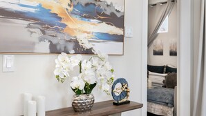 Et værelse med et træbord, hvor der står hvide lys, en vase med hvide blomster og en dekorativ tallerken. Et stort abstrakt maleri hænger på væggen over bordet.