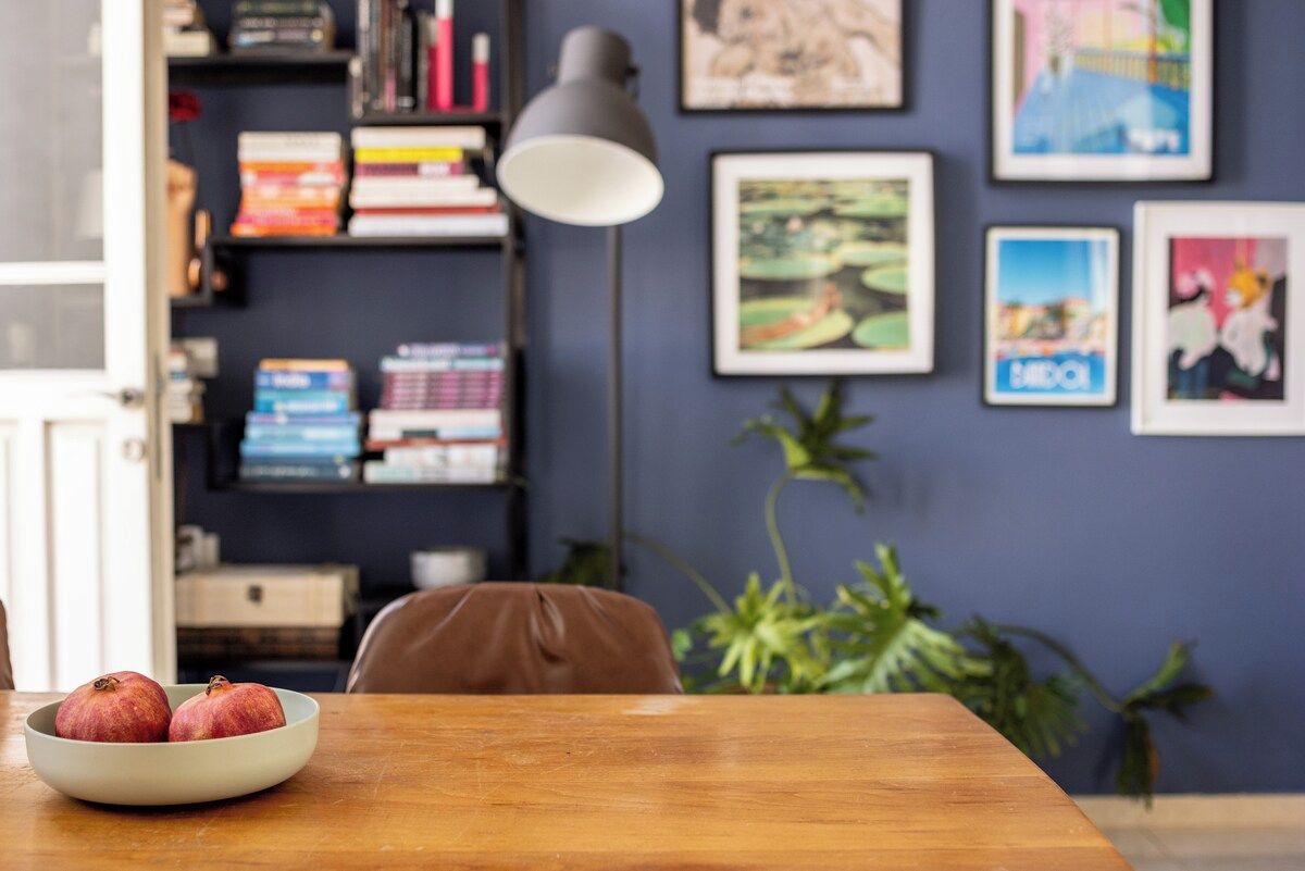 A room with a wooden table, a bowl of fruit, a bookshelf, and framed pictures on the wall.