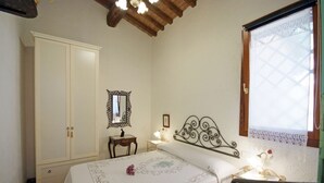 Een slaapkamer met een bed, een kledingkast, een nachtkastje met een lamp, een spiegel en een raam met kantgordijnen.