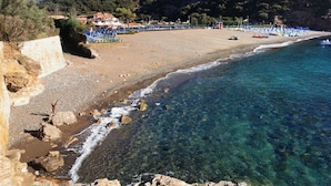 A beach with clear water, rocky shoreline, and buildings in the background.