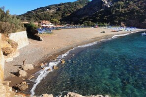 Una playa con agua cristalina, costa rocosa y edificios al fondo.