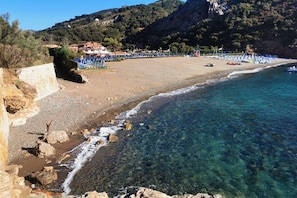 A beach with clear water, rocky shoreline, and buildings in the background.