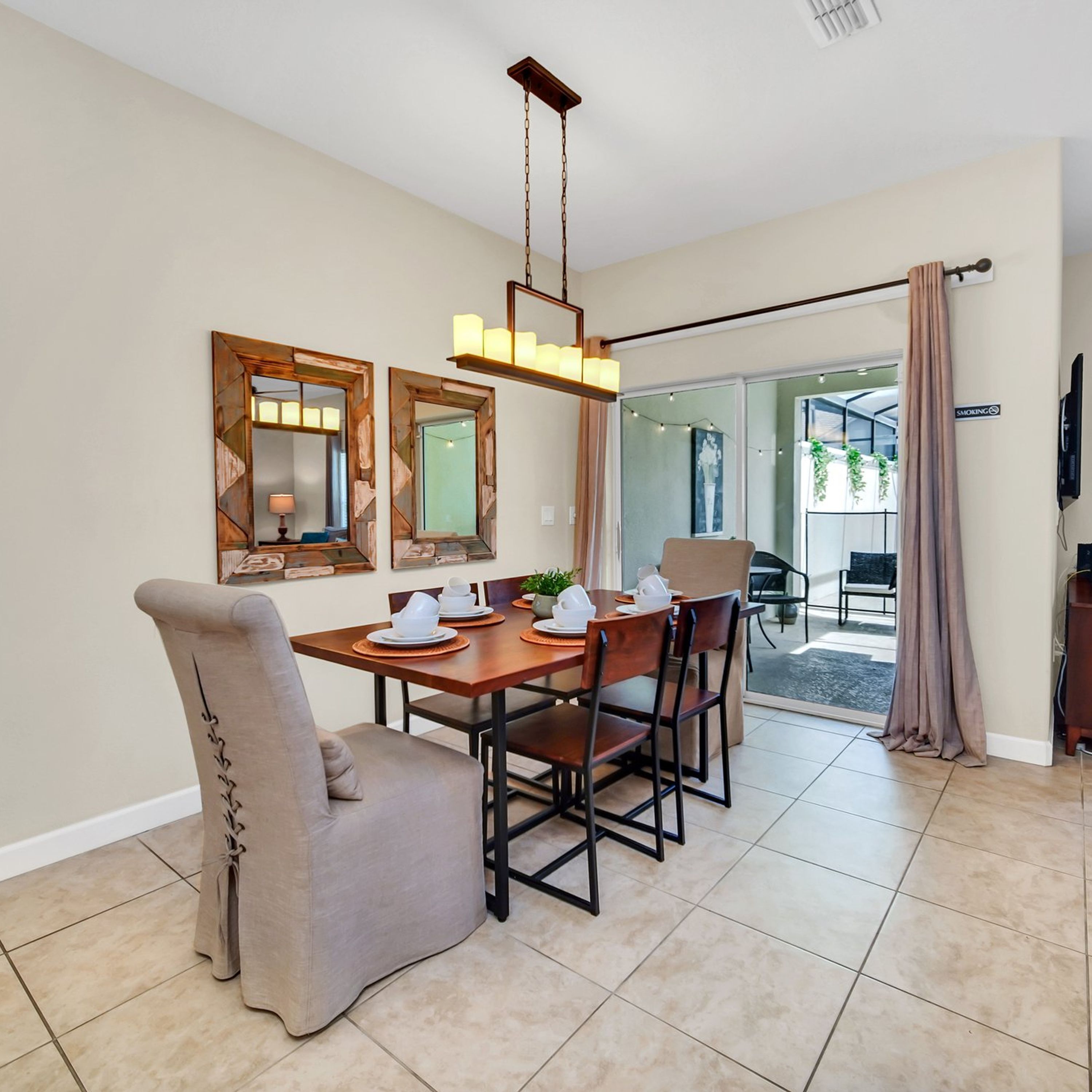 A dining area with a wooden table set for four, surrounded by chairs. There are two wall-mounted mirrors above the table, a hanging light fixture, and a sliding glass door leading to an outdoor area.