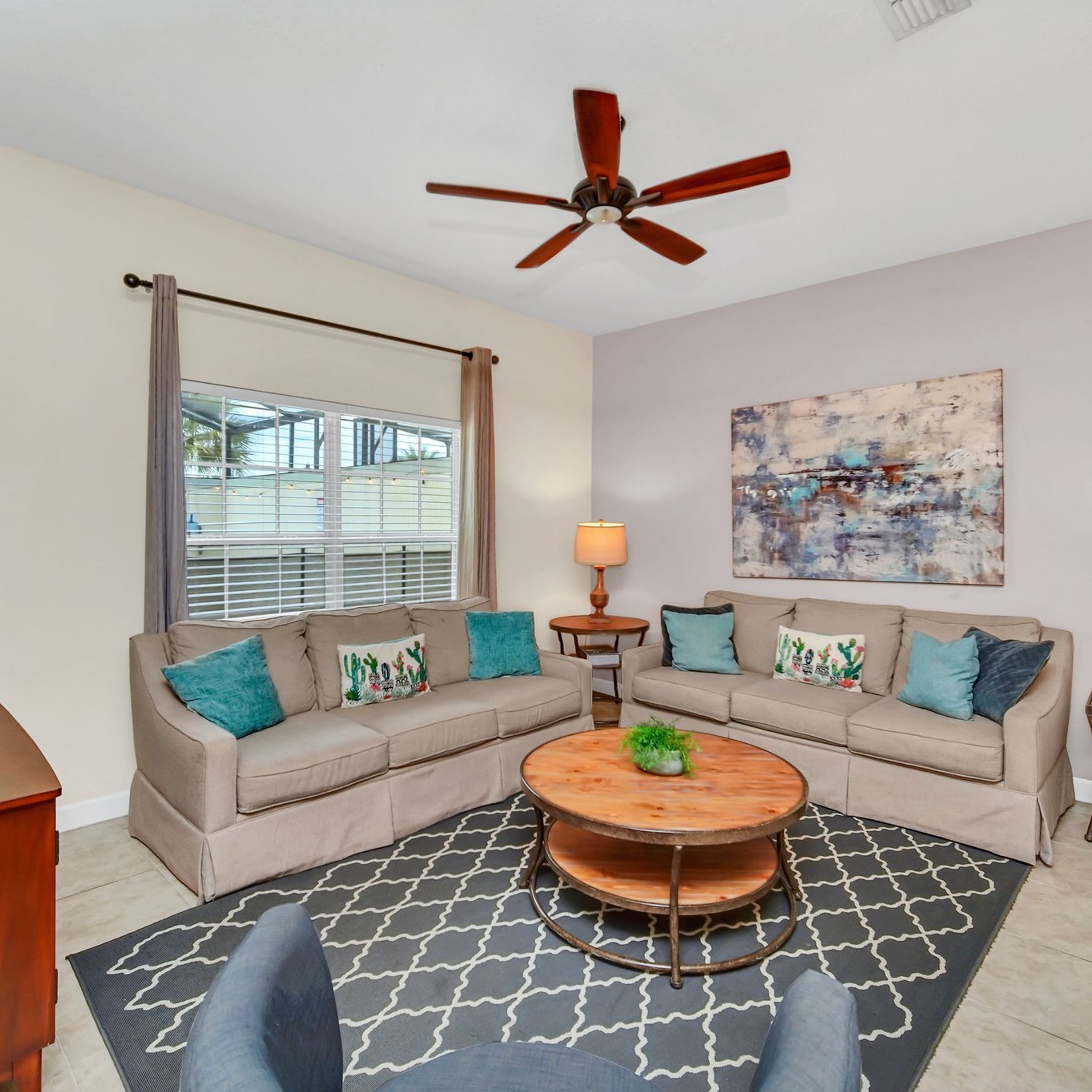 A living room with two beige sofas, a wooden coffee table, a ceiling fan, a window with blinds, and a painting on the wall.