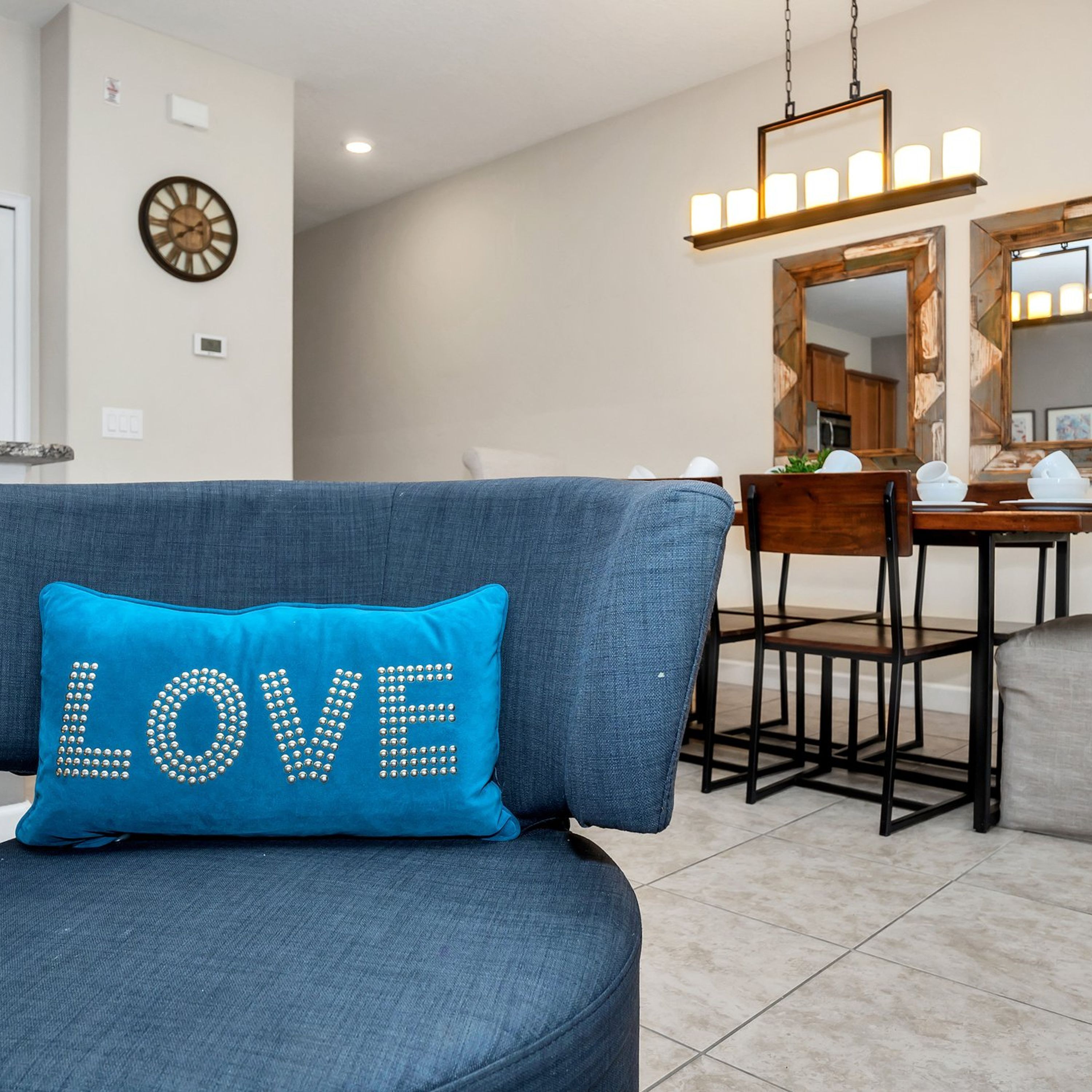 Ein Wohnzimmer mit einem blauen Sofa, einem Dekokissen mit der Aufschrift 'LOVE', einem Essbereich mit Holztisch und -stühlen sowie einer im Hintergrund sichtbaren Küche.
